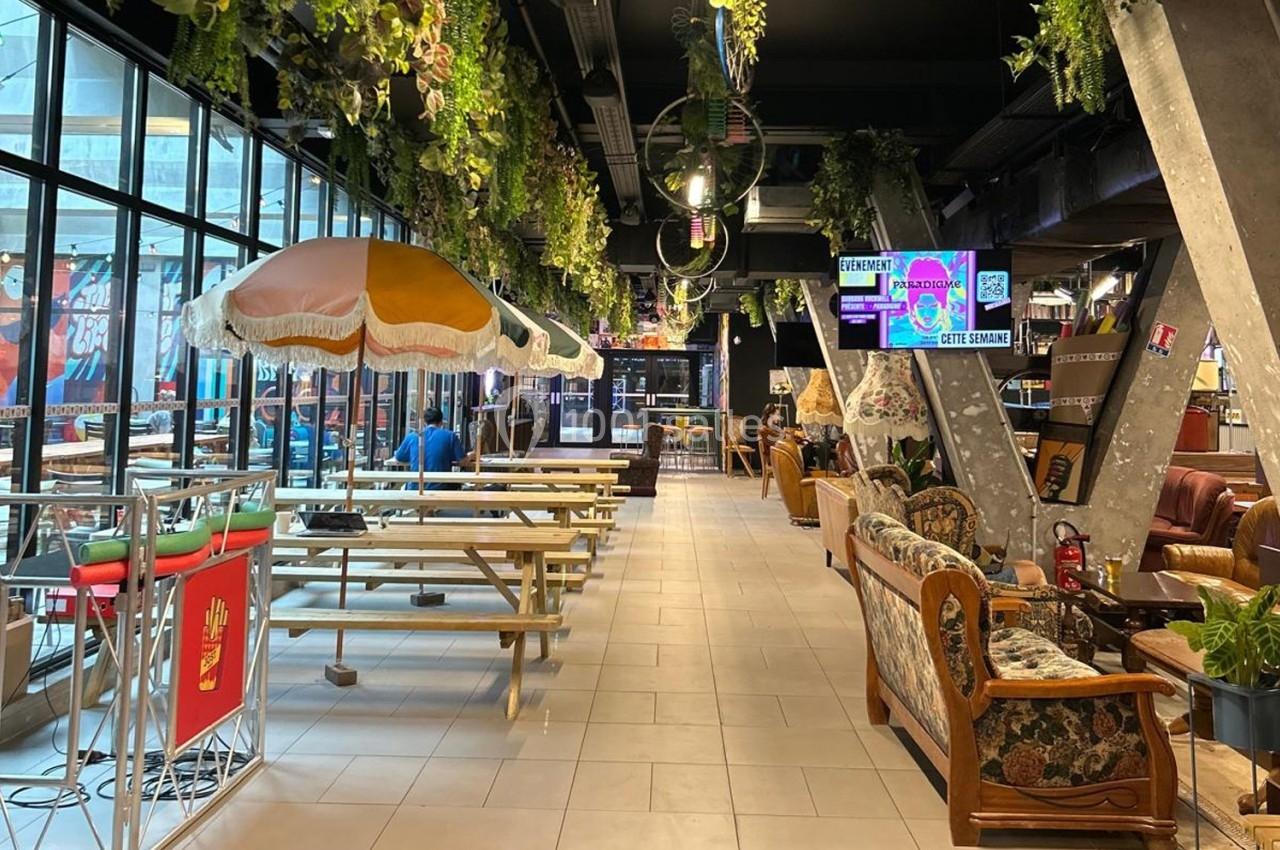Intérieur d'un espace de restauration avec tables en bois, parasols colorés et décorations végétales suspendues.