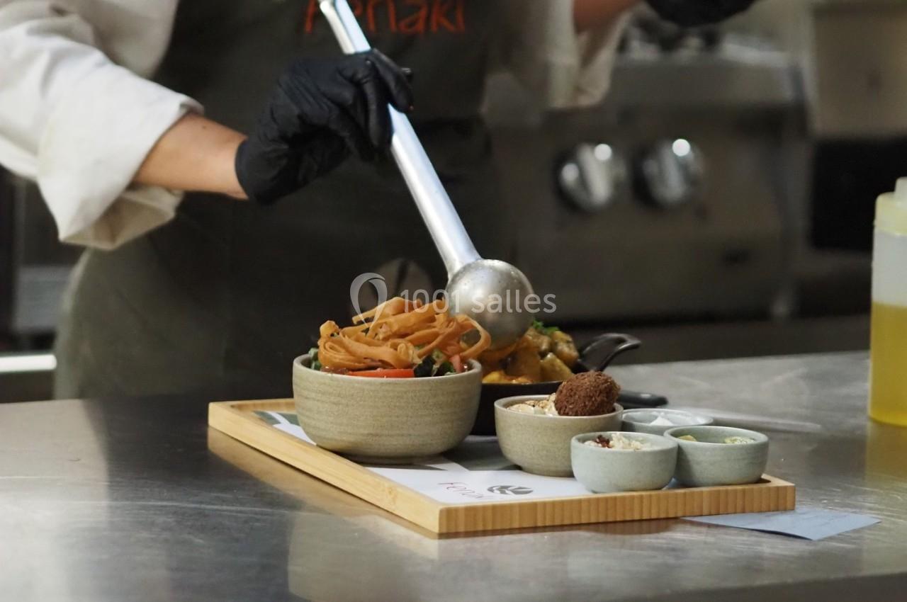 Une personne sert des nouilles dans un bol avec une louche, sur un plateau contenant divers accompagnements.