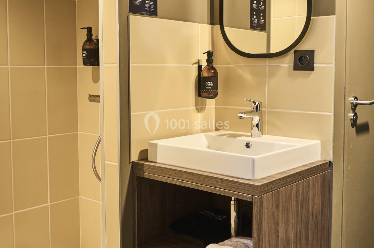 Lavabo moderne avec meuble en bois, miroir ovale, distributeur de savon mural et carrelage beige.