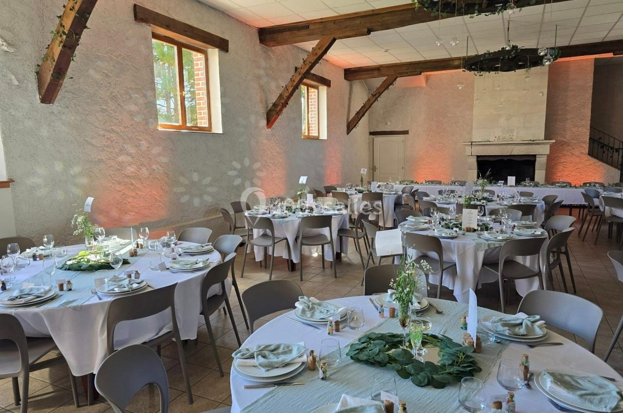 Salle de réception décorée pour un événement, avec tables rondes dressées, nappes blanches et éléments végétaux.