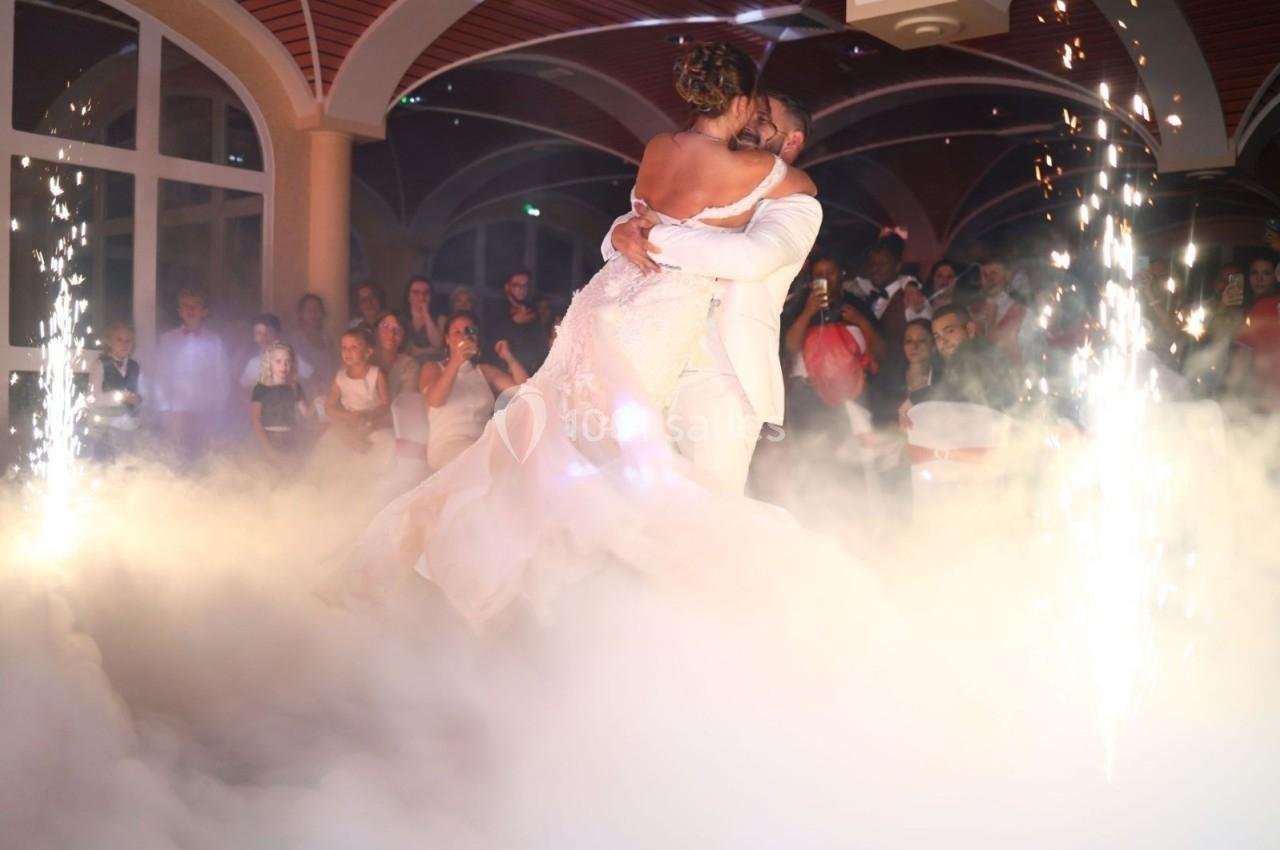 Un couple de mariés danse au milieu de la fumée et des étincelles, entouré d'invités dans une salle festive.