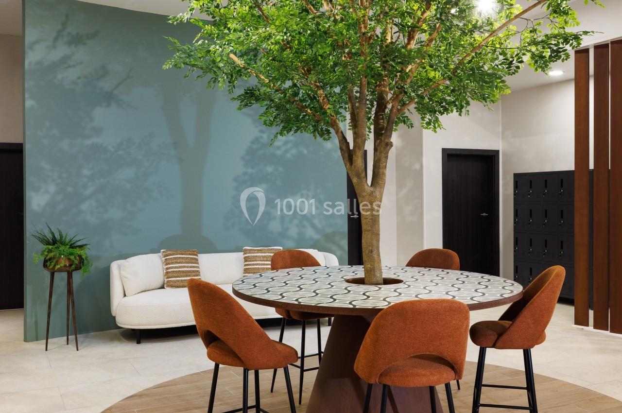 Table ronde avec un arbre en son centre, entourée de chaises orange, dans un espace intérieur moderne avec un mur bleu.