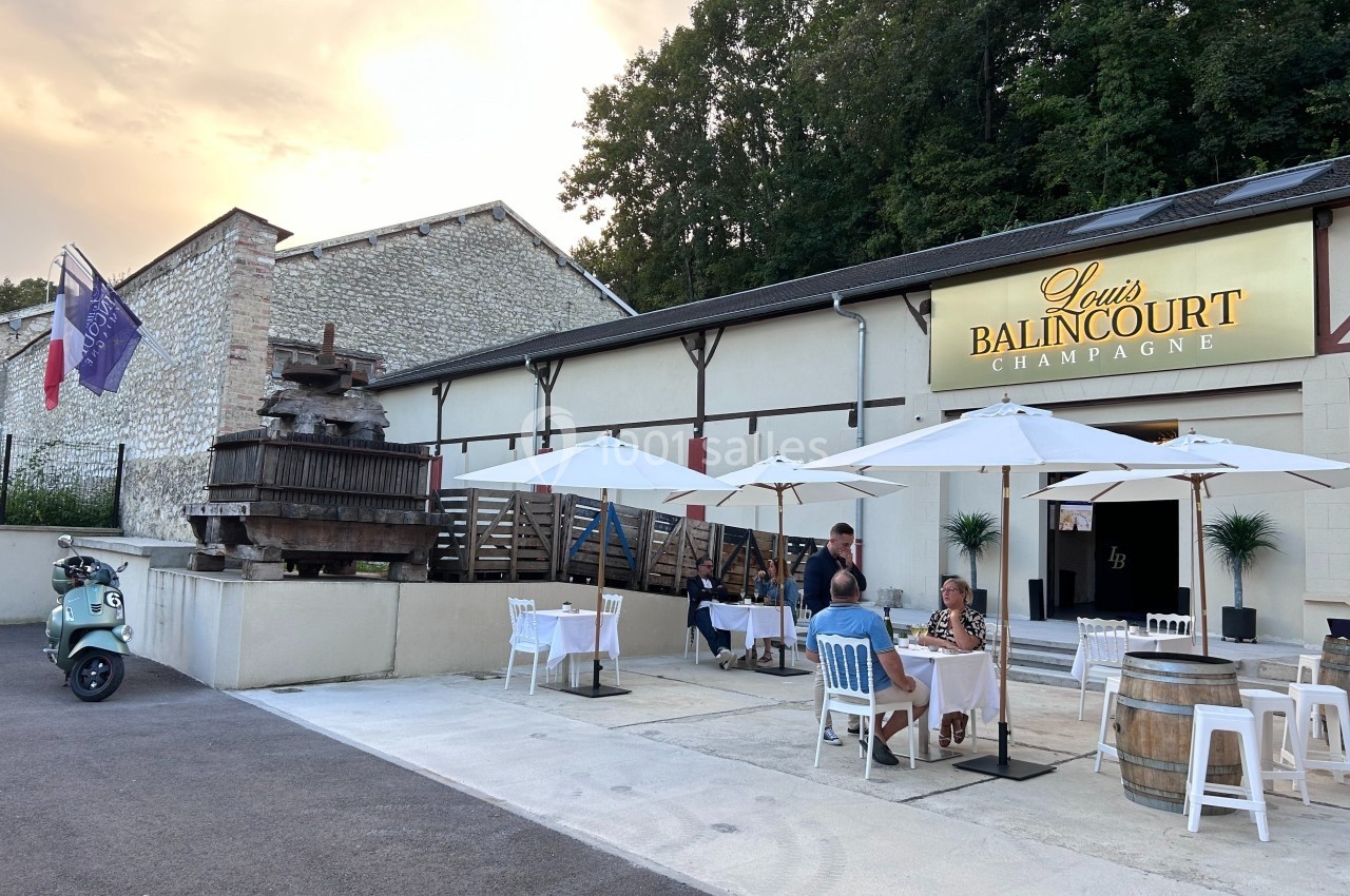 Façade d'une maison de champagne avec terrasse aménagée, tables sous parasols et ancien pressoir en bois.