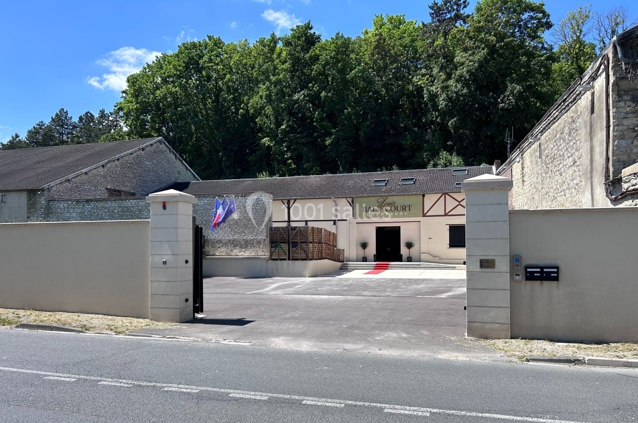 Entrée d'un bâtiment avec façade en pierre, drapeaux français, portail ouvert et forêt en arrière-plan.