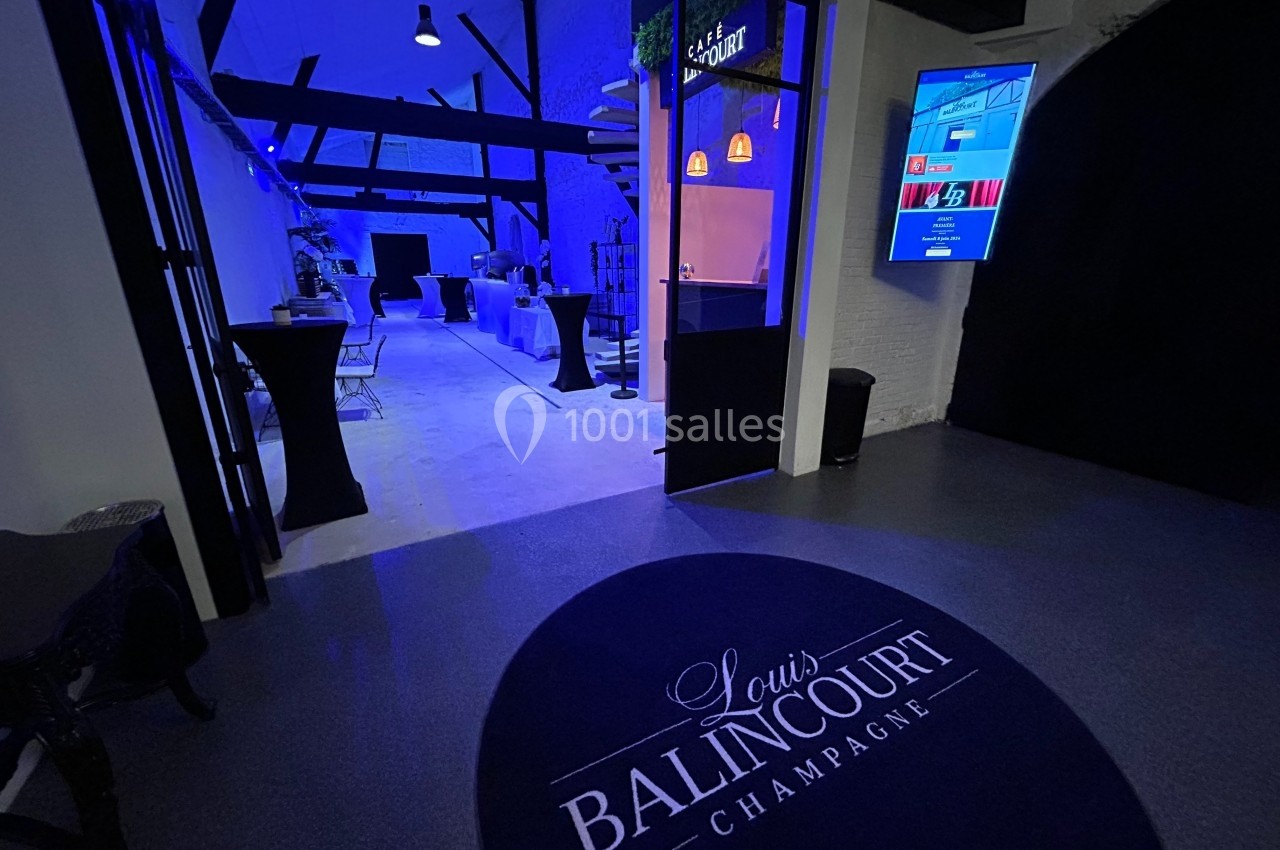 Entrée d'une salle éclairée en bleu avec tables hautes noires, logo ’Louis Balincourt Champagne’ au sol.