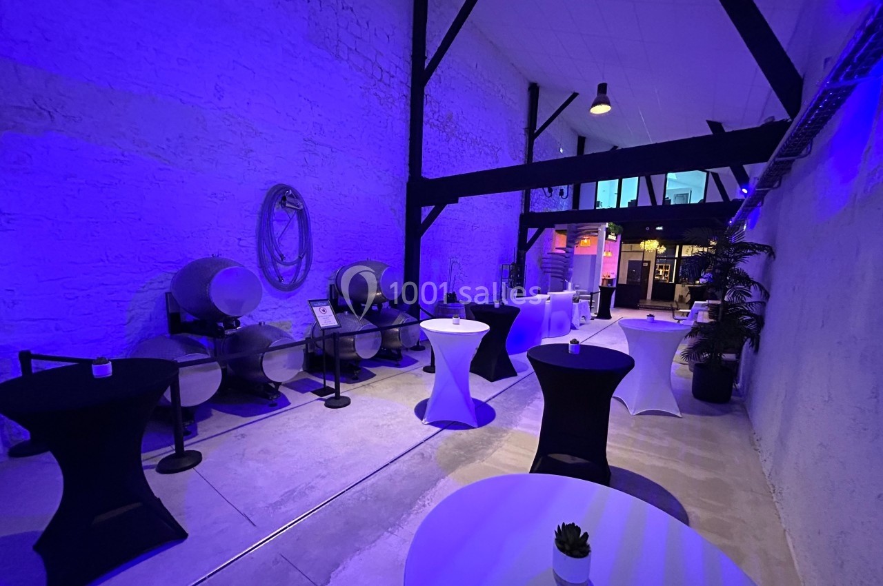 Salle avec murs en pierre, barriques de vin alignées, tables hautes drapées et éclairage bleu tamisé.