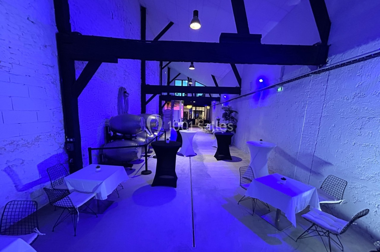 Salle avec murs blancs, poutres noires apparentes, éclairage bleu, tables dressées et chaises modernes.