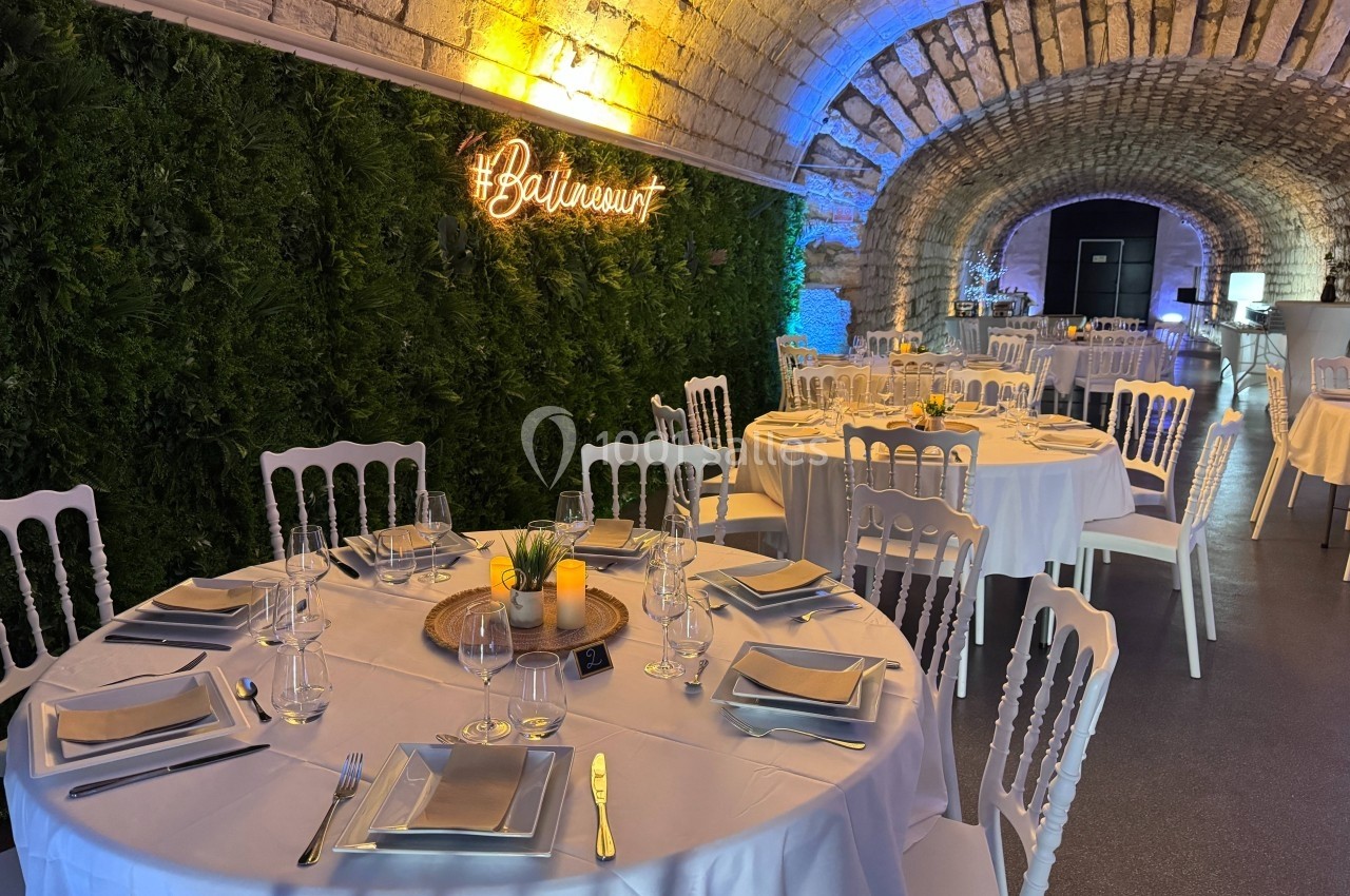 Salle voûtée décorée avec des tables rondes dressées pour un événement, éclairage chaleureux et mur végétal.