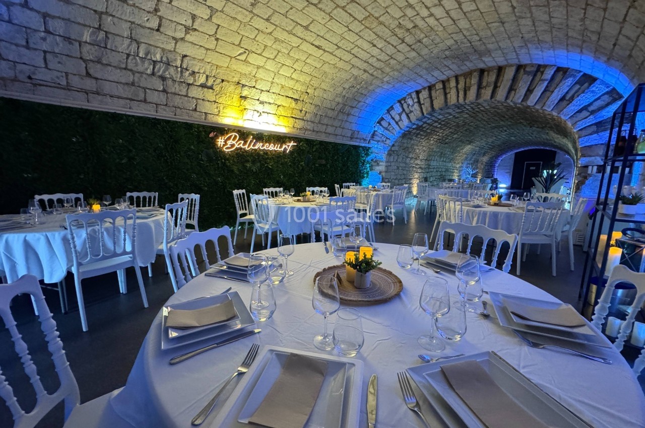 Salle voûtée décorée pour un événement, avec tables rondes dressées et éclairage tamisé bleu et jaune.