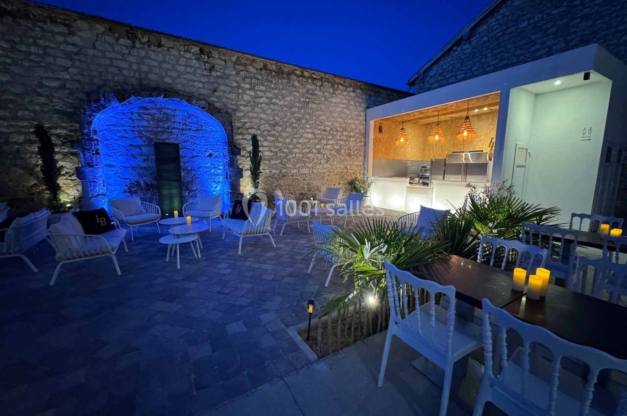 Cour extérieure éclairée en soirée, avec des tables, des chaises, des plantes et un espace cuisine moderne.