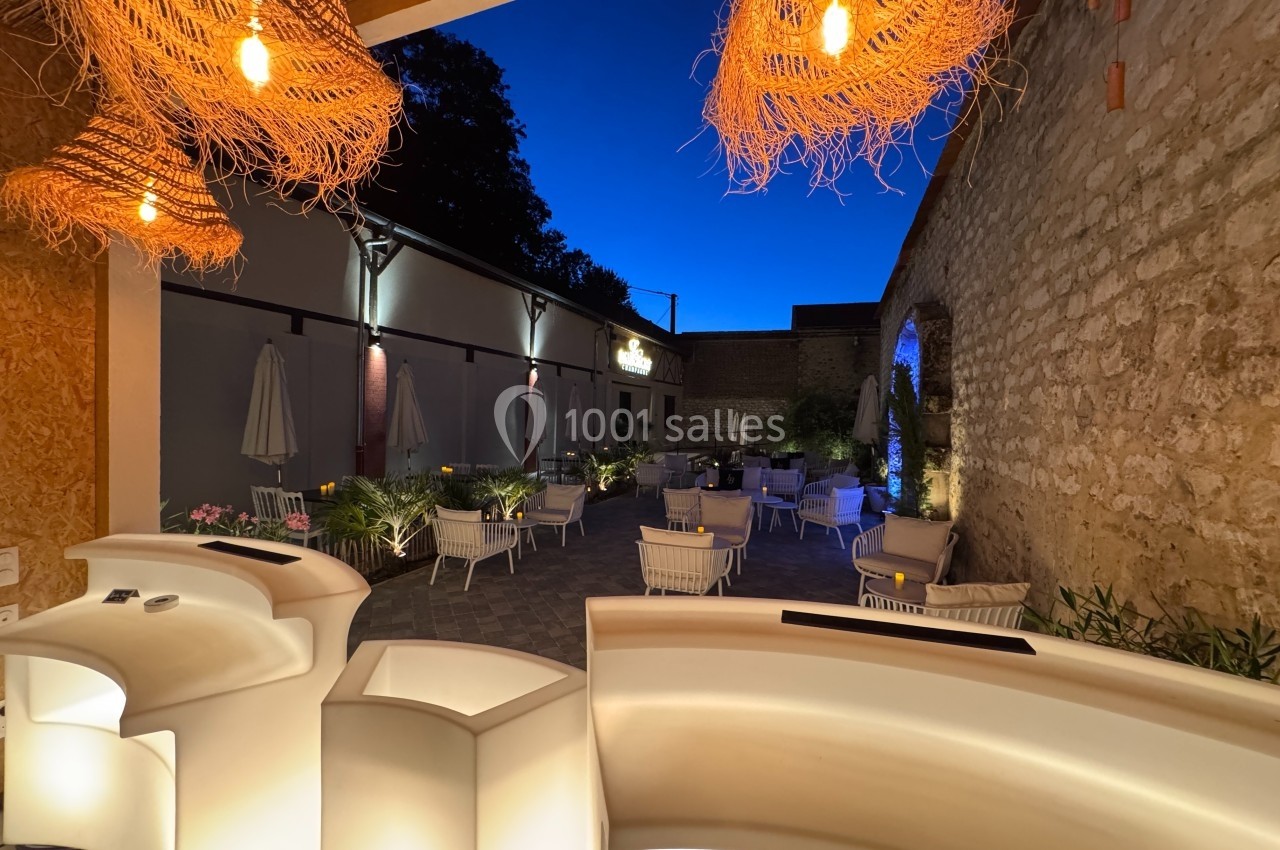 Terrasse extérieure éclairée en soirée, avec des tables, des chaises blanches et des lampes suspendues en osier.