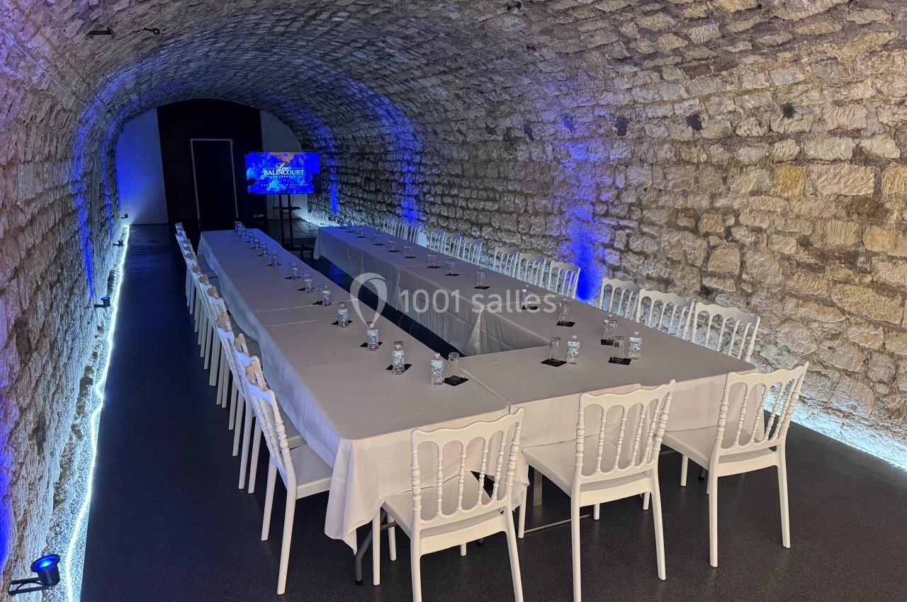 Salle voûtée en pierre aménagée avec deux longues tables dressées, chaises blanches et éclairage bleu tamisé.