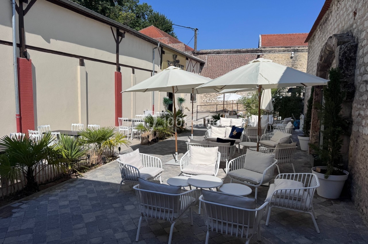 Terrasse extérieure pavée avec chaises blanches, tables et parasols, entourée de murs en pierre et végétation.