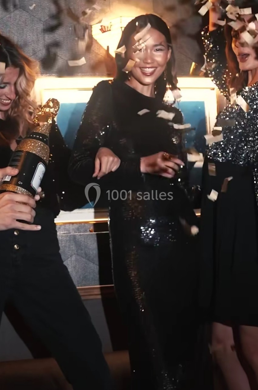 Trois femmes en tenue de soirée célèbrent sous des confettis, l'une tenant une bouteille décorative.