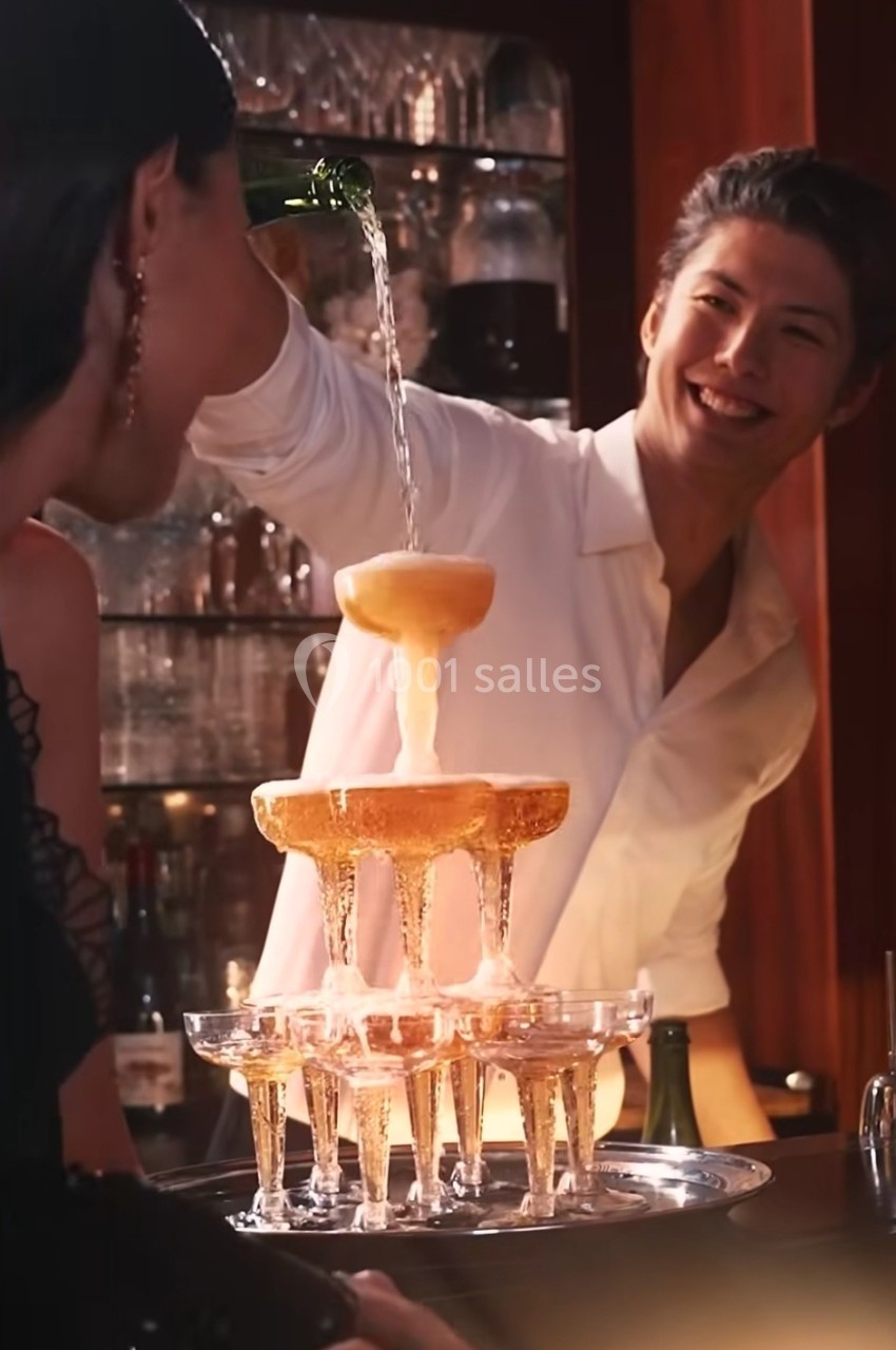 Un homme verse du champagne dans une pyramide de coupes, entouré de convives dans un bar élégant.