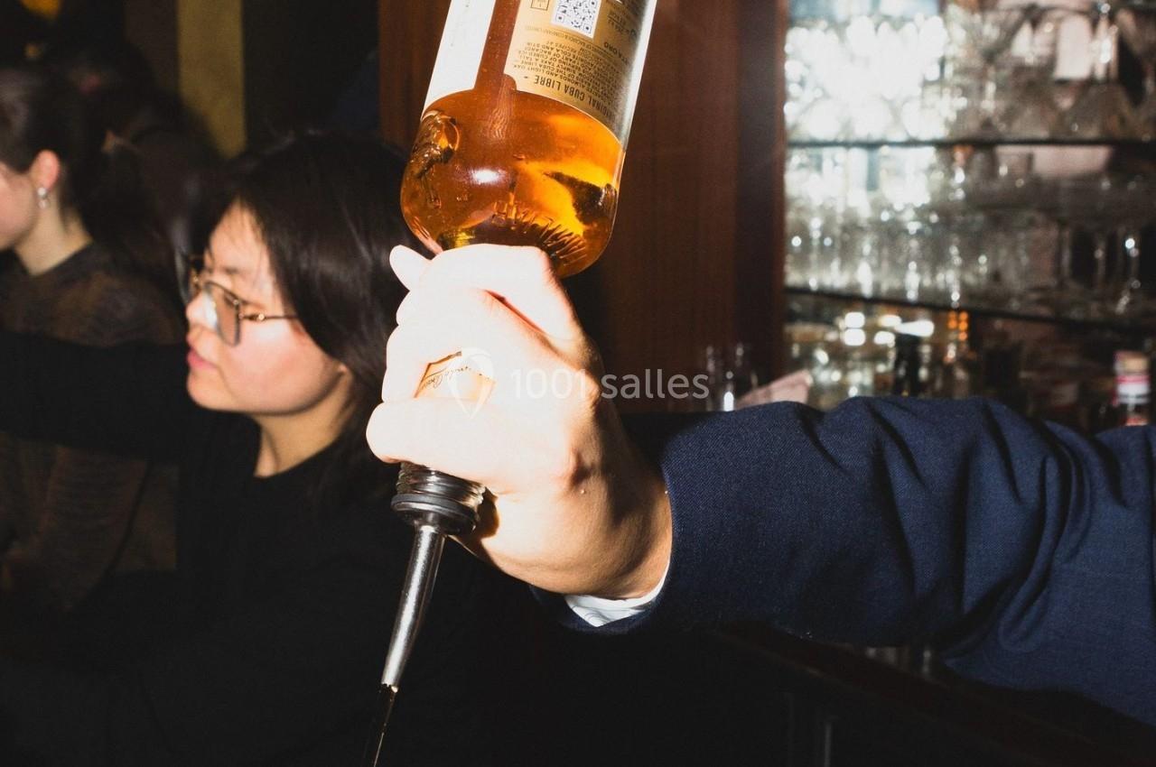Une main verse une boisson alcoolisée depuis une bouteille dans un bar, avec des verres en arrière-plan.