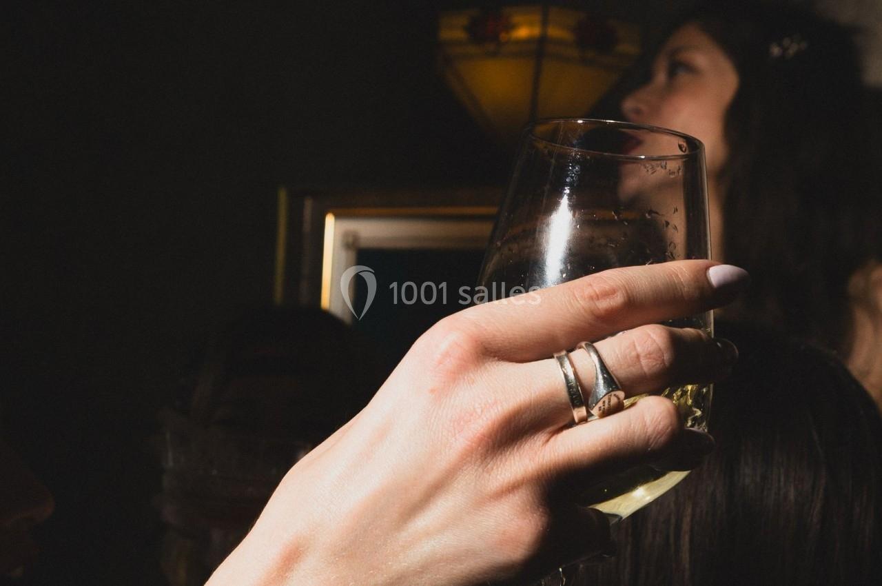 Main tenant un verre de vin blanc dans un environnement sombre avec des personnes en arrière-plan flou.