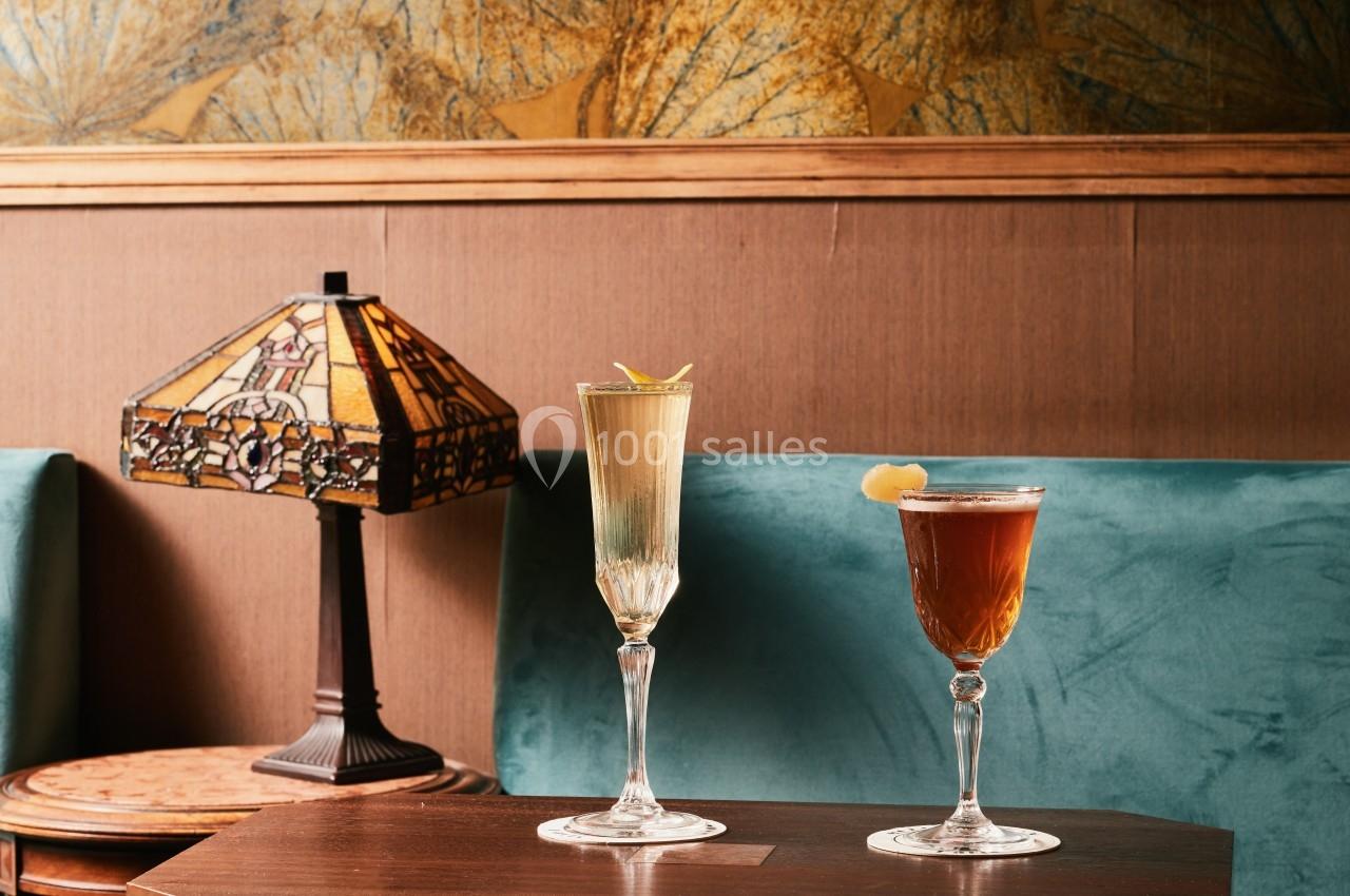 Deux verres de cocktails élégants posés sur une table en bois, avec une lampe vintage et un canapé en velours vert en…