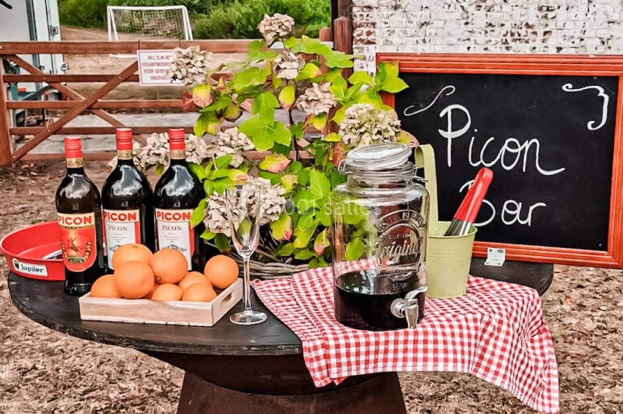 Table avec des bouteilles de Picon, des oranges, un verre, une carafe et une ardoise indiquant ’Picon Bar’.