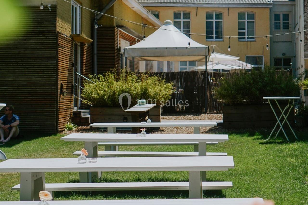 Tables de pique-nique blanches alignées sur une pelouse, entourées de bâtiments et d'un espace ombragé avec des parasols.