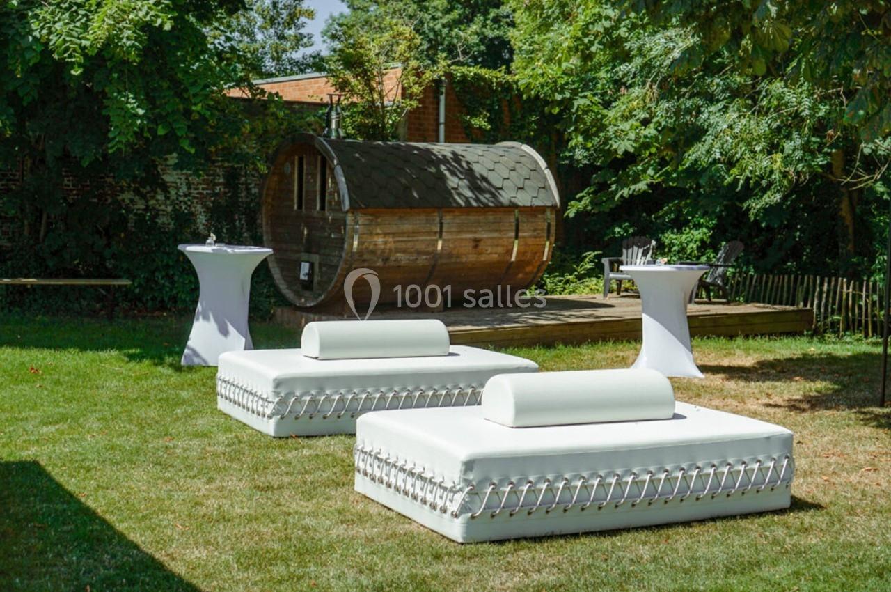 Deux lits de jardin blancs sur une pelouse, avec une cabane en bois et des tables hautes en arrière-plan.