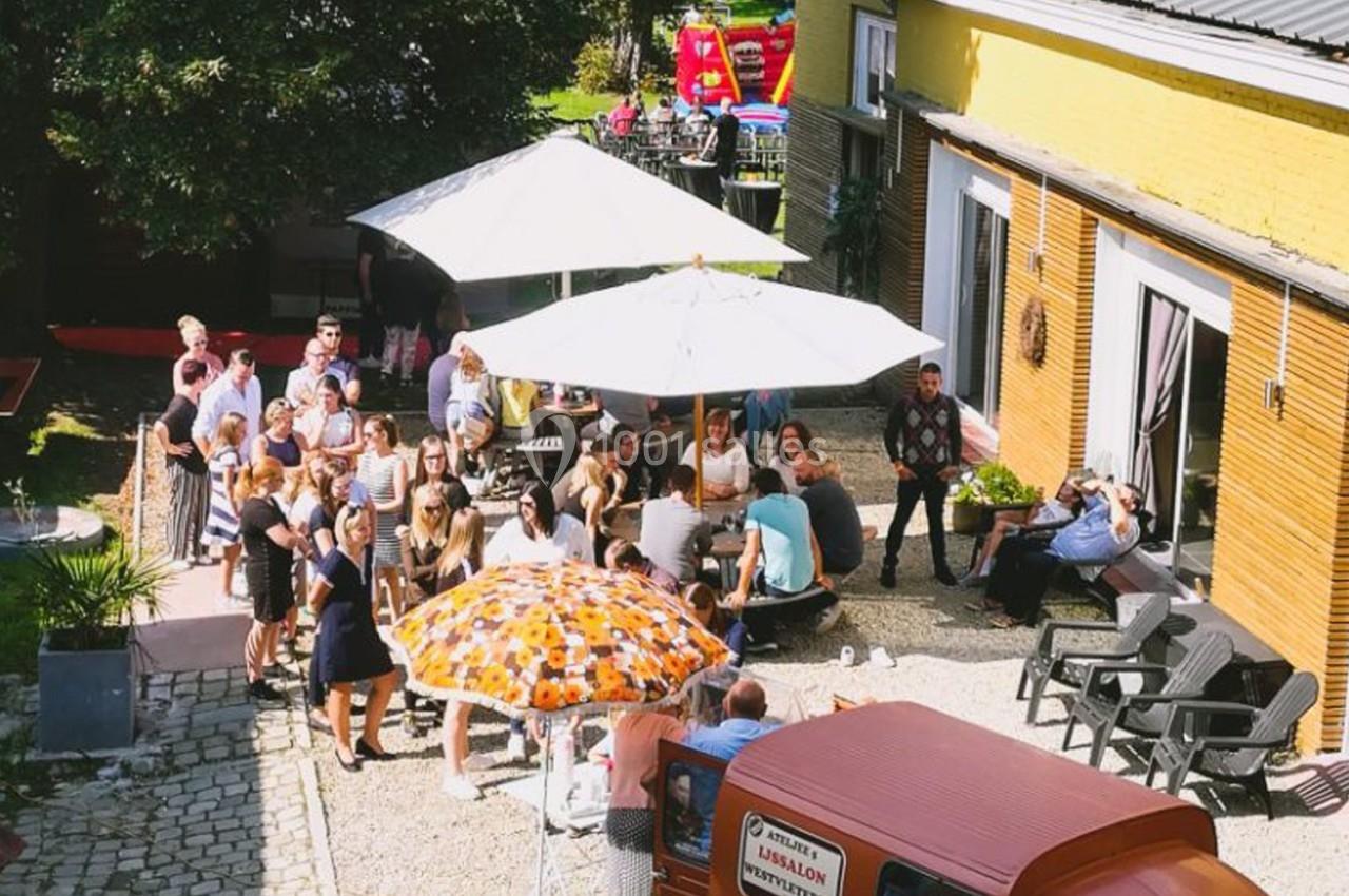 Groupe de personnes réunies dans une cour extérieure ensoleillée, avec parasols et mobilier de jardin.