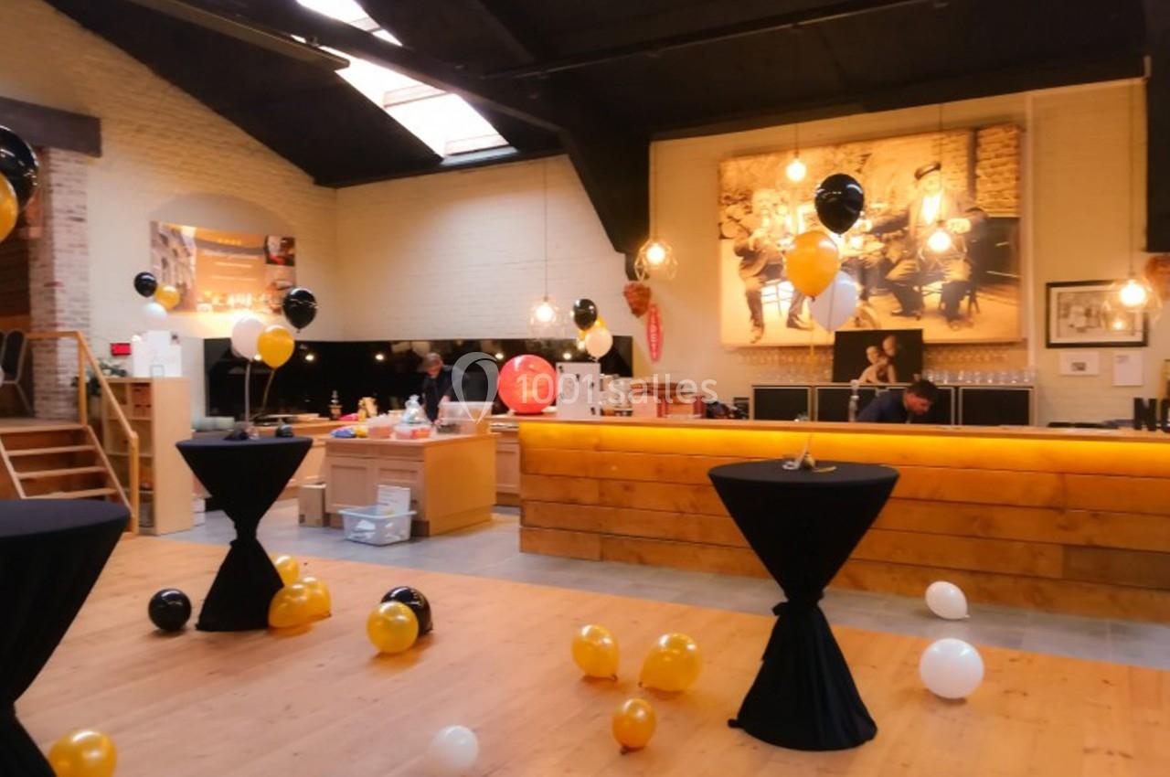 Salle décorée pour un événement avec ballons noirs, blancs et jaunes, tables hautes et éclairage chaleureux.