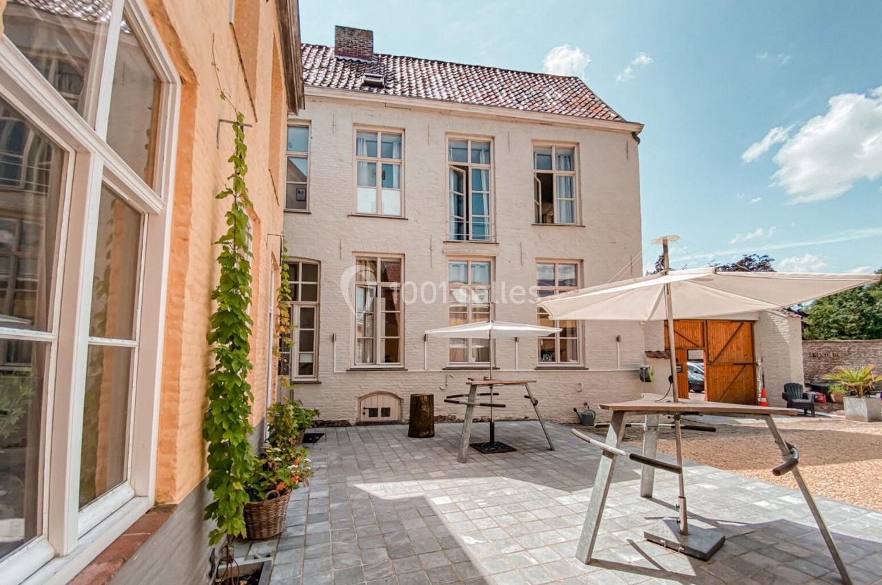 Cour pavée avec mobilier de jardin, parasols blancs et façade en briques claires sous un ciel dégagé.
