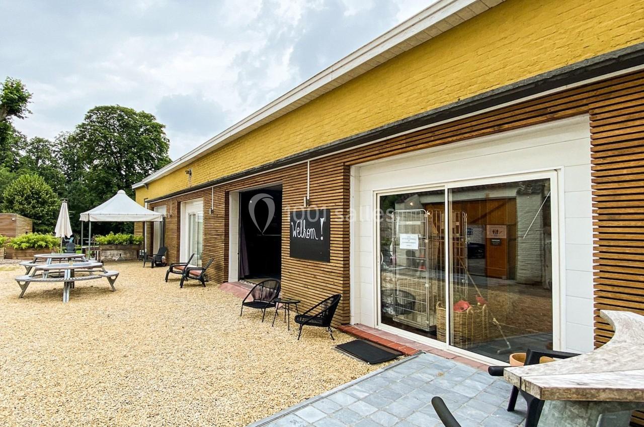 Façade d'un bâtiment jaune avec terrasse en gravier, tables de pique-nique et chaises devant une entrée vitrée.