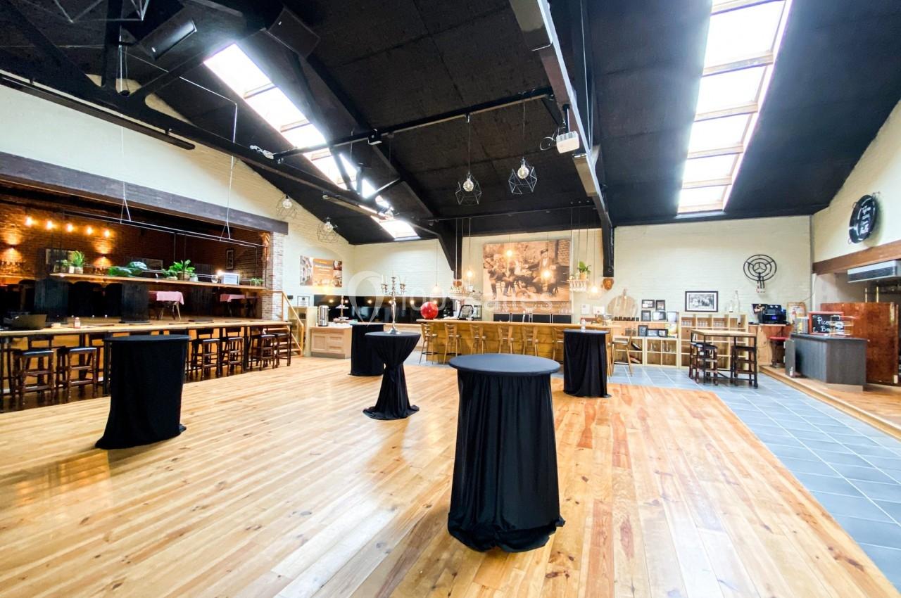 Salle spacieuse avec parquet en bois, tables hautes couvertes de nappes noires et lumière naturelle provenant de verrières.