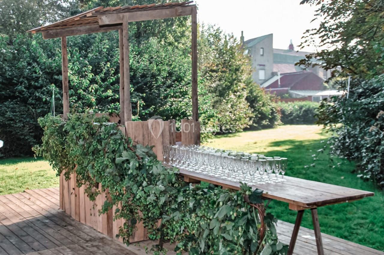 Un bar en bois décoré de feuillage, installé en extérieur sur une terrasse, avec des rangées de verres alignés.