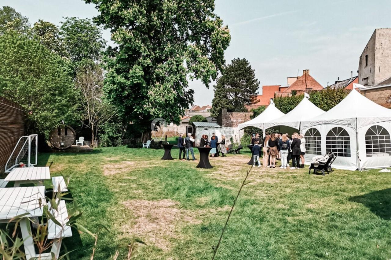 Jardin avec pelouse, arbres, tentes blanches et un groupe de personnes rassemblées près de tables hautes.