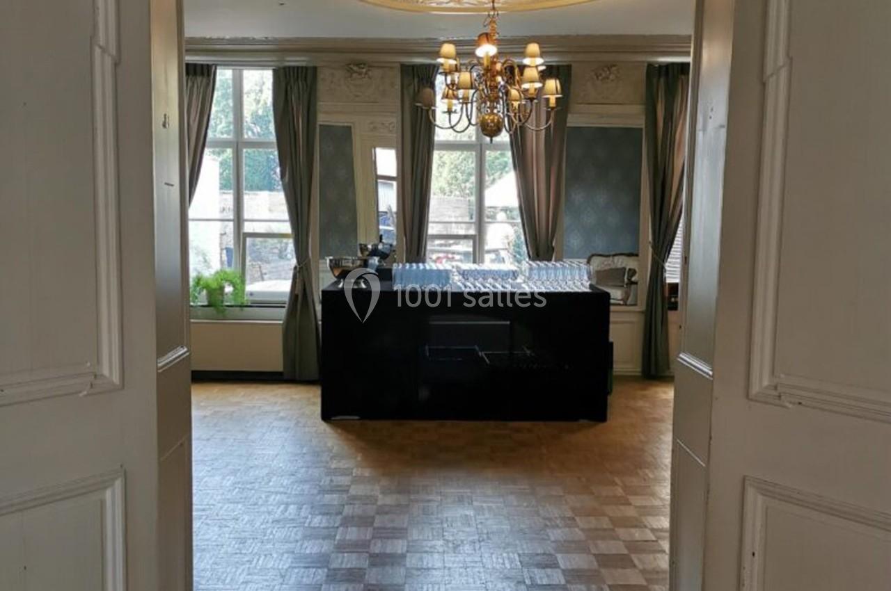 Salle lumineuse avec parquet, table couverte de verres, grandes fenêtres, rideaux et lustre suspendu.