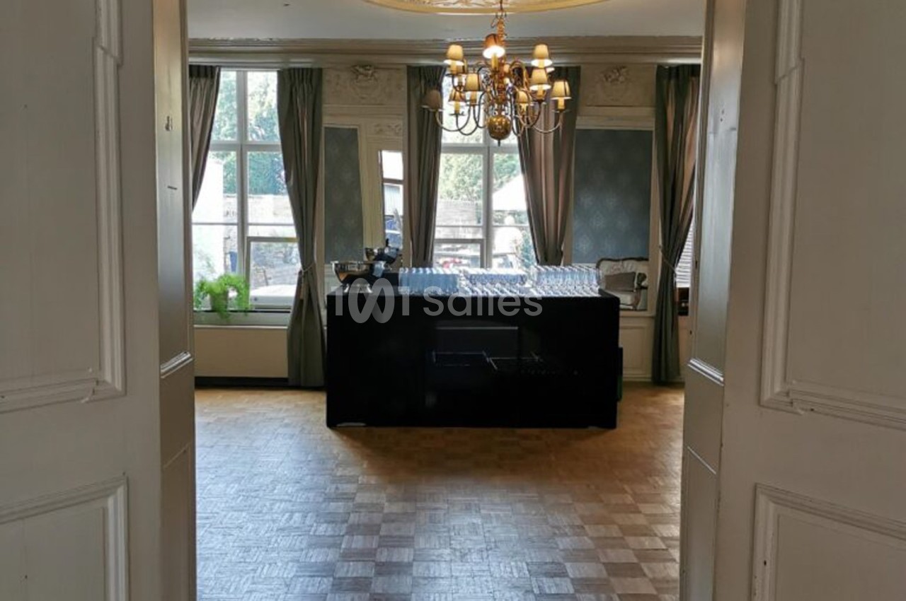 Salle lumineuse avec parquet, table couverte de verres, grandes fenêtres, rideaux et lustre suspendu.