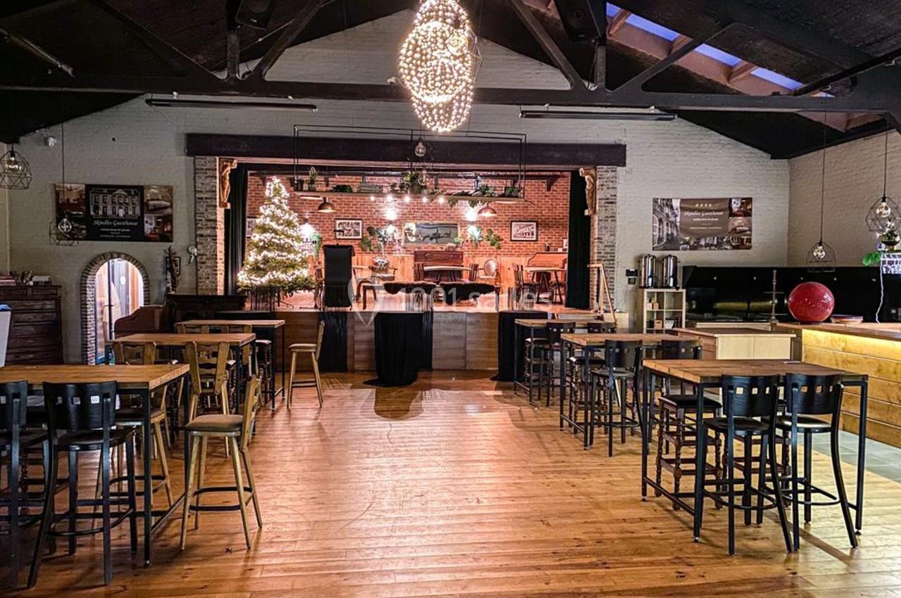 Salle avec parquet en bois, tables hautes, scène décorée pour Noël et éclairage suspendu.