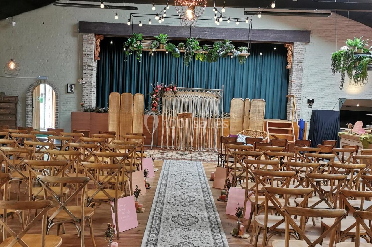 Salle de réception décorée pour une cérémonie, avec des chaises en bois alignées et une arche florale devant une scène.