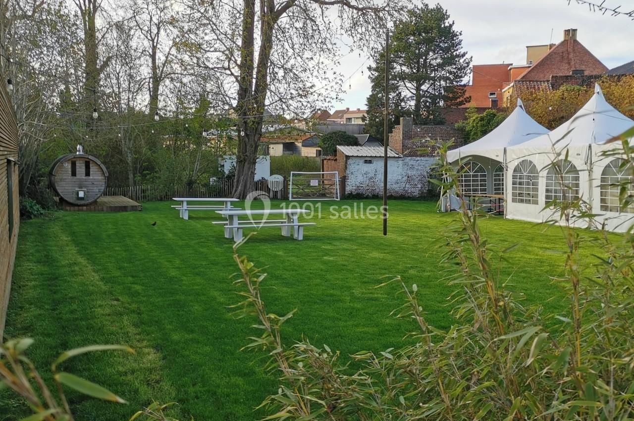 Jardin avec pelouse, tables blanches, tente blanche, tonneau en bois et arbres en arrière-plan sous un ciel dégagé.