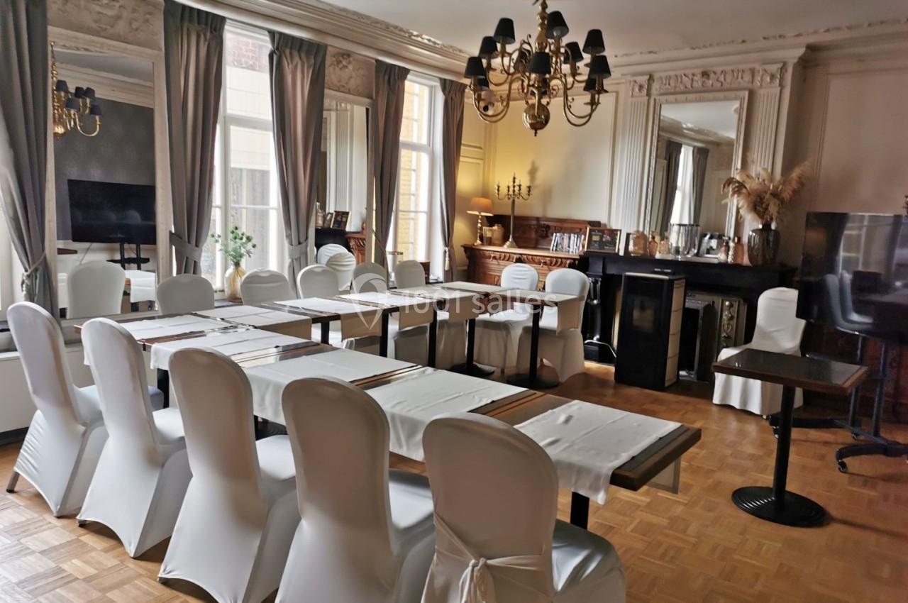 Salle élégante avec tables alignées, chaises blanches, lustre, grandes fenêtres et décoration classique.