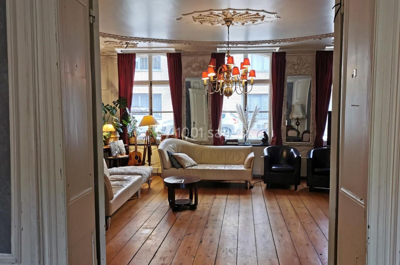 Salon lumineux avec parquet en bois, canapés, fauteuils, lustre et grandes fenêtres encadrées de rideaux rouges.