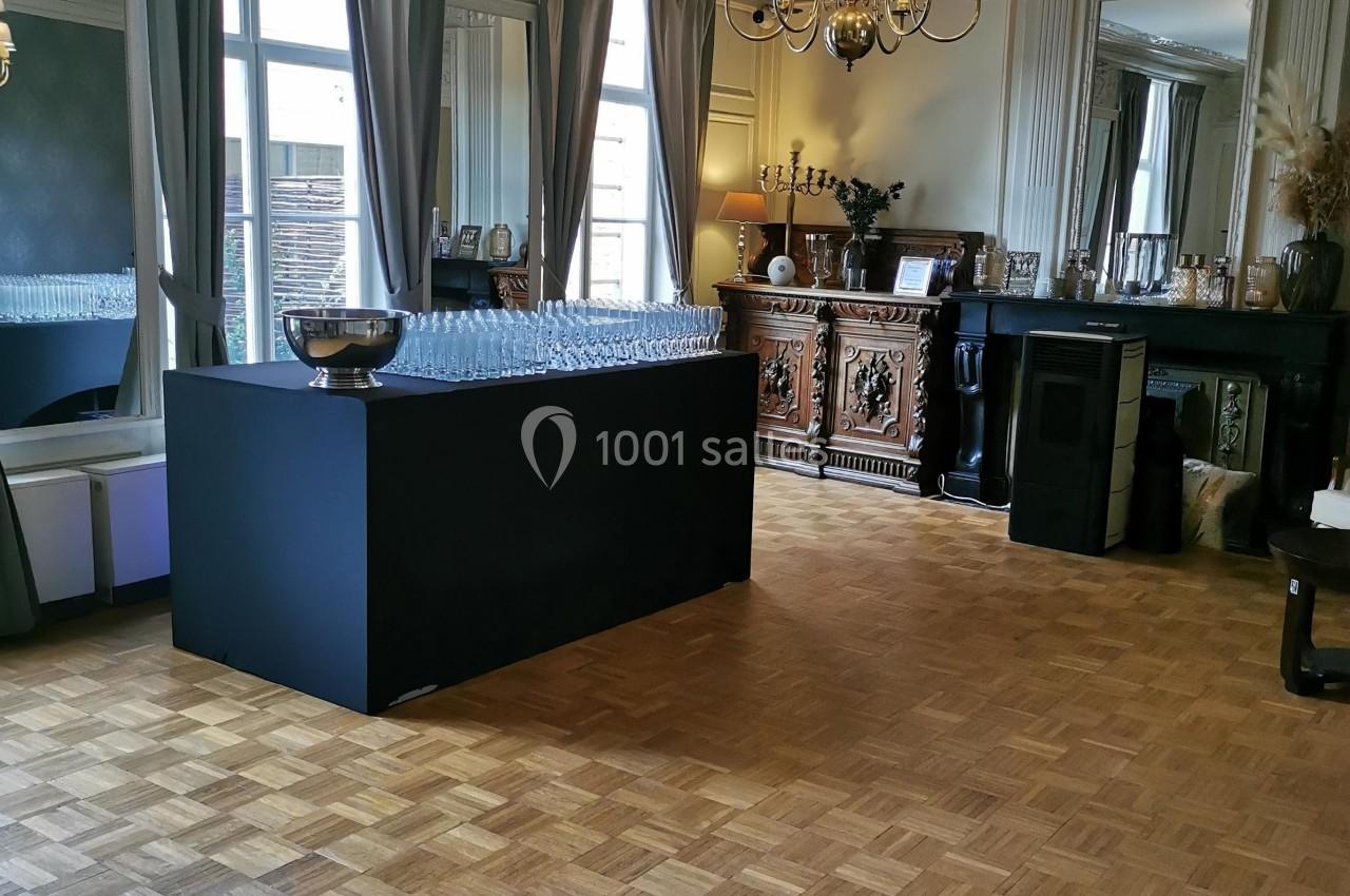 Salle élégante avec parquet, comptoir noir, lustre central et grandes fenêtres encadrées de rideaux.