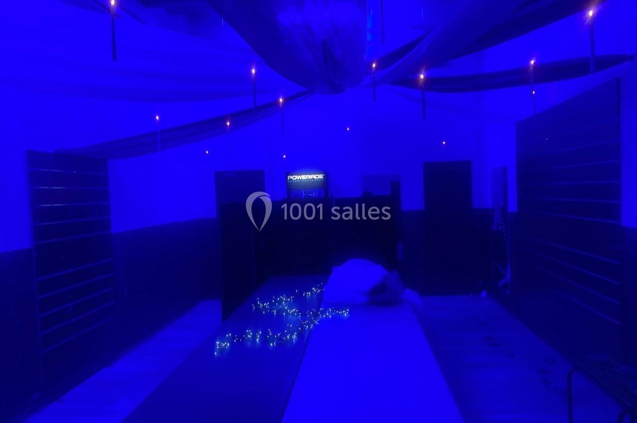 Salle sombre éclairée par une lumière bleue, avec des tables alignées et des guirlandes lumineuses au centre.