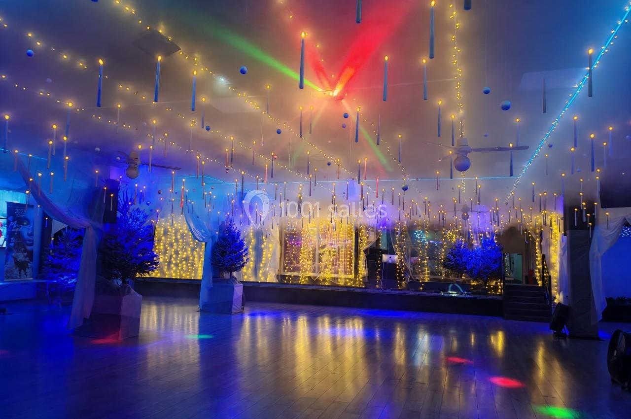 Salle décorée de guirlandes lumineuses suspendues, éclairée par des projecteurs colorés et entourée de sapins illuminés.