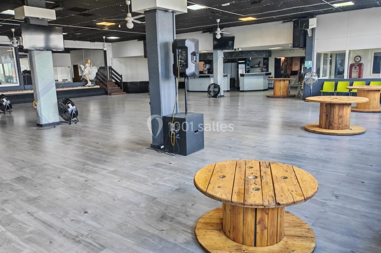 Salle spacieuse avec parquet gris, colonnes, tables en bois rondes et une scène en arrière-plan.