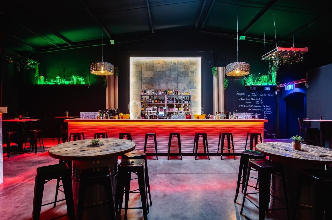 Bar moderne avec comptoir éclairé en rouge, tabourets noirs, tables en bois et décoration végétale sous un éclairage tamisé.