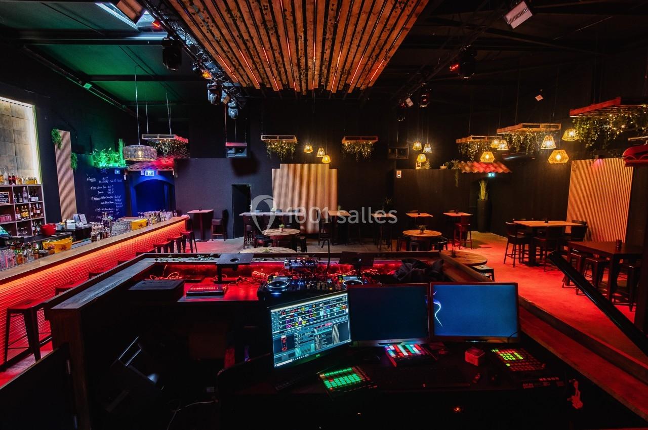 Salle de bar avec éclairage tamisé, tables en bois, bar à gauche et espace DJ avec équipements électroniques au premier plan.