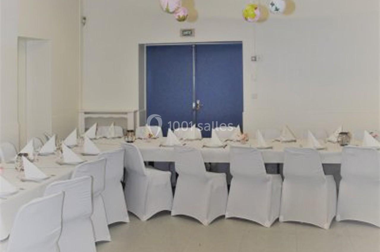 Salle décorée avec des tables alignées, nappes blanches, chaises houssées et lampions suspendus au plafond.