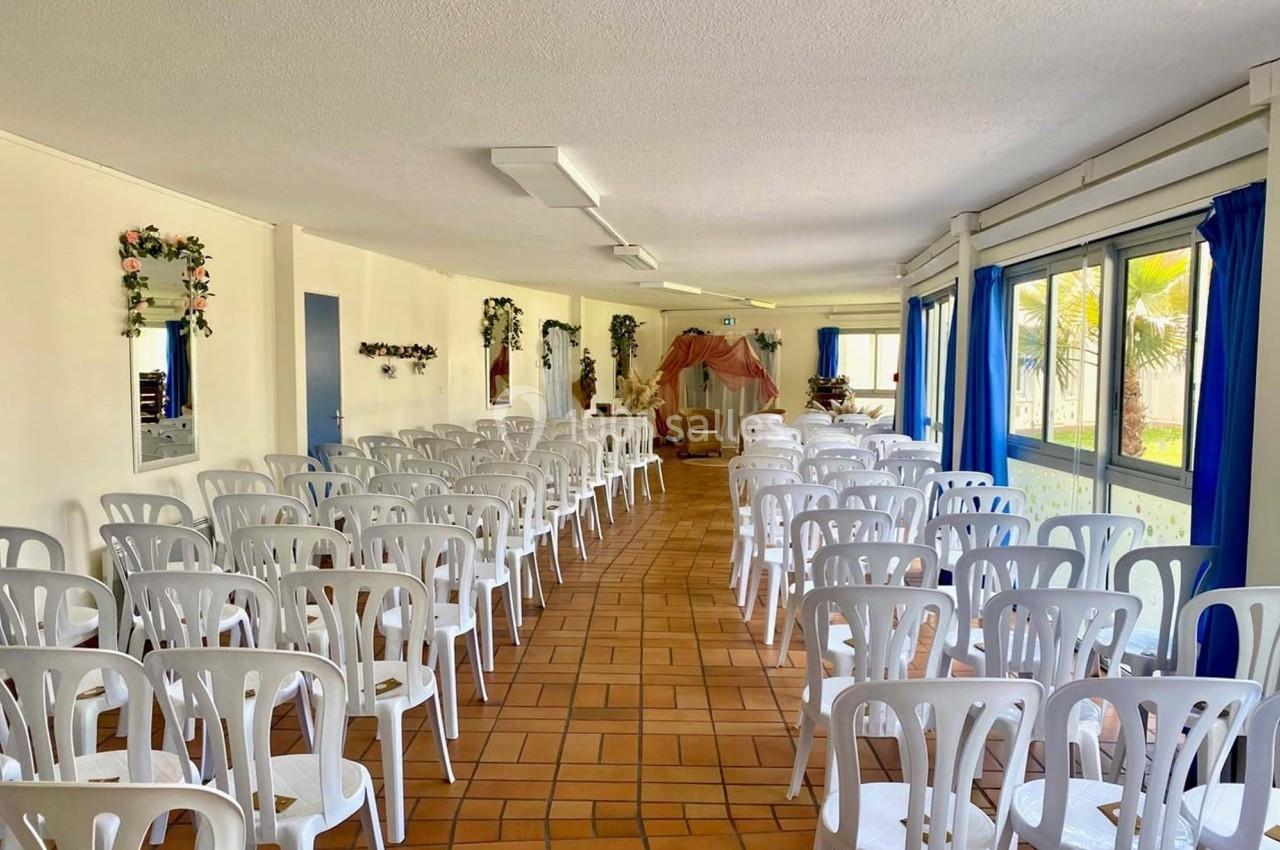 Salle lumineuse aménagée avec des rangées de chaises blanches, décorée de fleurs et ouverte sur l'extérieur par de grandes…