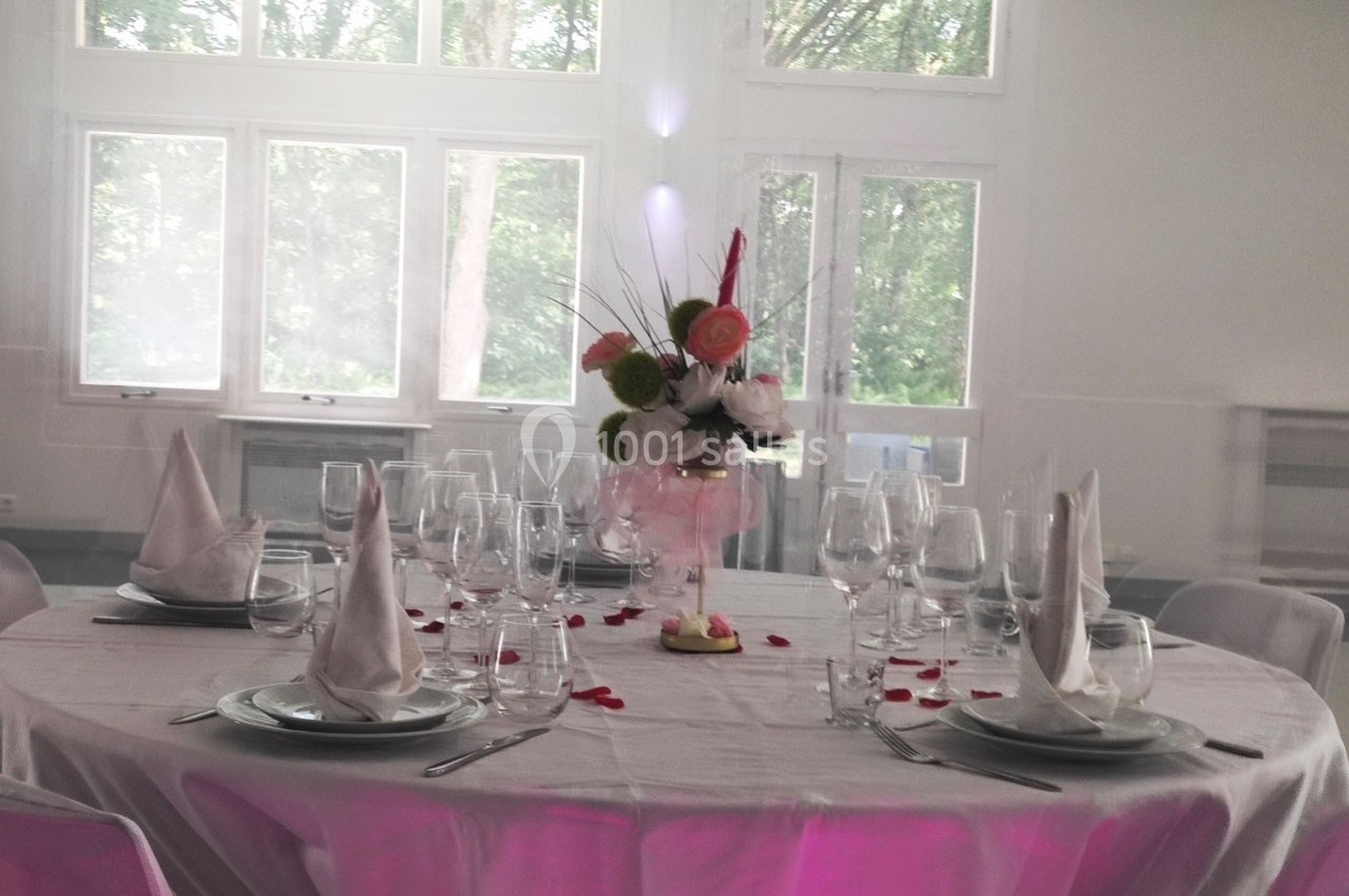 Table ronde dressée avec une nappe blanche, verres, serviettes pliées et un centre de table floral dans une salle lumineuse.