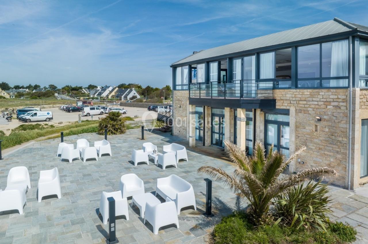 Terrasse en pierre avec mobilier blanc devant un bâtiment moderne en bord de mer, sous un ciel dégagé.