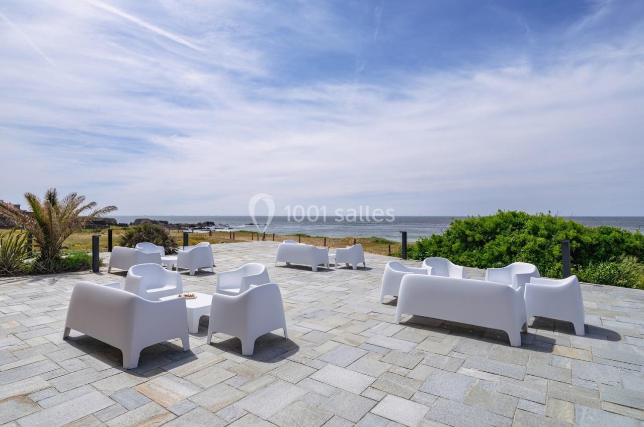 Terrasse en pierre avec mobilier blanc moderne, vue sur la mer et végétation côtière sous un ciel partiellement nuageux.